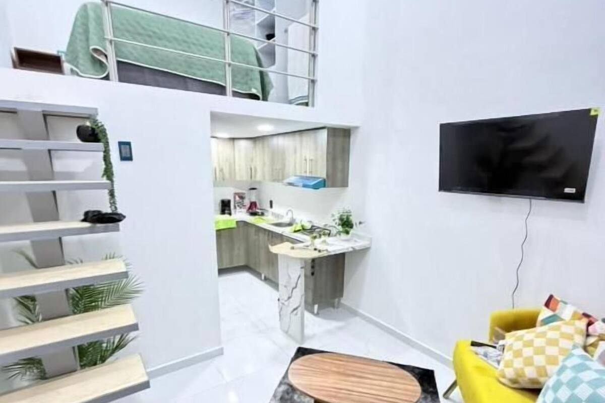 A modern apartment with a kitchen, a dining area, a living room with a yellow sofa, and a TV mounted on the wall.
