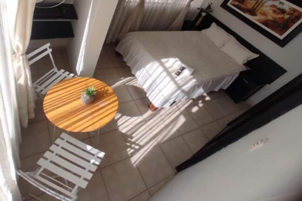 A small, compact room with a bed, a round table with a plant, a chair, a fan, and a painting on the wall.