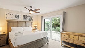 Un dormitorio con una cama grande, un ventilador de techo, una mesita de noche con lámpara, un aparador de madera y una puerta corrediza de vidrio que da a un jardín.