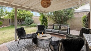 Un patio techado con techo blanco, lámpara de mimbre, sillas negras, un sofá gris y una mesita redonda pequeña.
