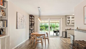 Un comedor con mesa y sillas de madera, una estantería, una lámpara colgante y vistas al exterior a través de un ventanal.