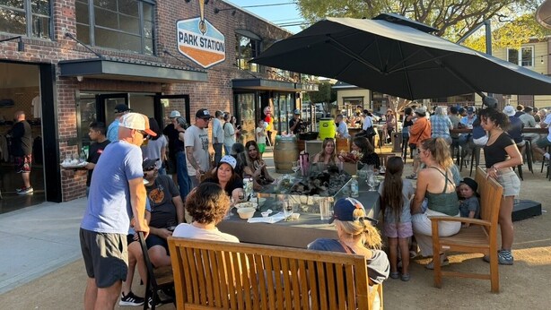 Un espace extérieur aménagé pour les repas, avec des tables et des chaises, un bâtiment en briques sur lequel on peut lire l’inscription « PARK STATION », et des personnes rassemblées autour.