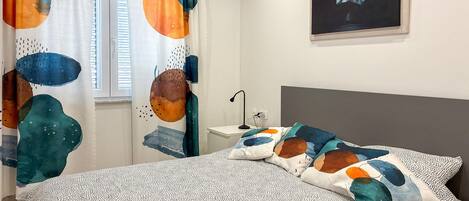A bedroom with a bed, pillows, a nightstand, a lamp, a window with curtains, and a painting on the wall.