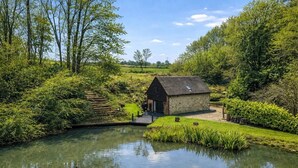 A rustic stone building with a sloped roof, surrounded by lush greenery and a calm pond. - Lake House Staycation (Hollingbourne)