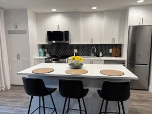 Cocina moderna con armarios blancos, refrigerador de acero inoxidable, microondas e isla central con tres taburetes negros.