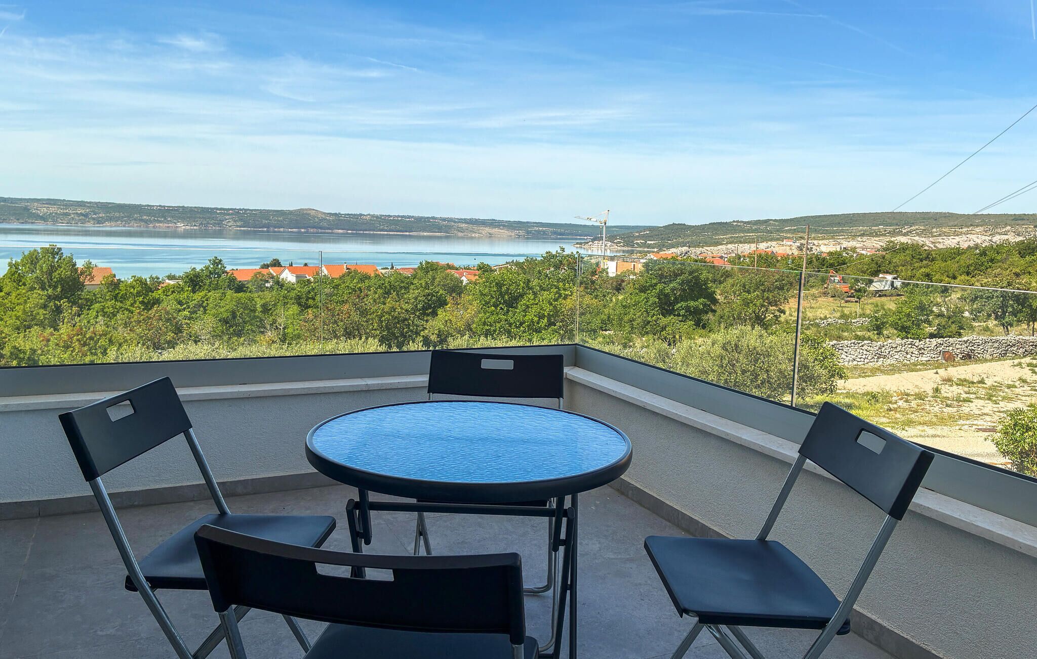 Un balcon avec une table ronde et quatre chaises, donnant sur un paysage côtier avec des bâtiments et des arbres.