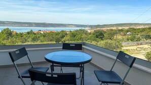 Varanda com mesa redonda e quatro cadeiras, com vista para paisagem costeira com edifícios e árvores.