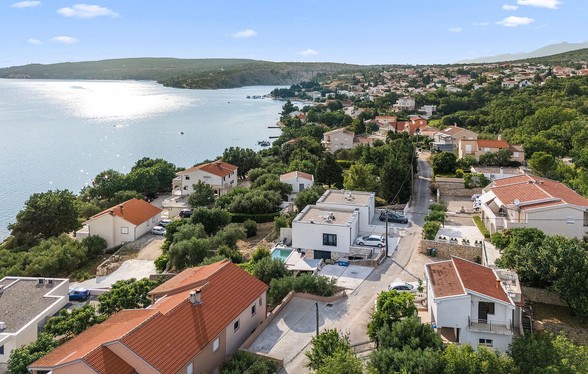 A coastal residential area with houses, a clear view of the sea, and a hilly landscape in the background.