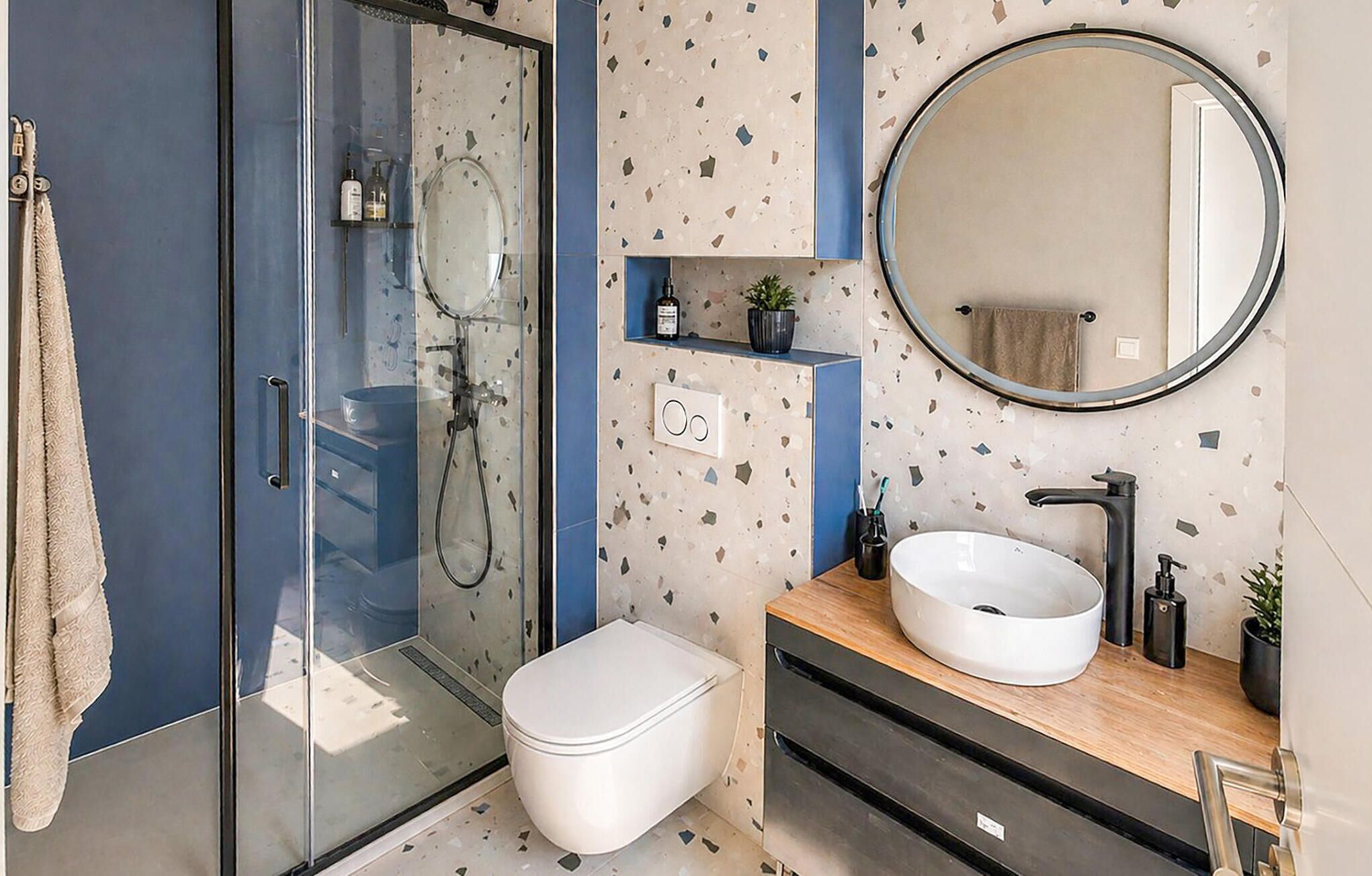 A modern bathroom with a glass shower enclosure, a white toilet, a round mirror, and a sink with a wooden countertop.
