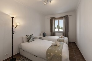 A hotel room with two beds, a standing lamp, a window with curtains, and a tiled floor.