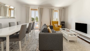 A modern living room with a dining table, sofa, TV, and a large window with curtains. - Casa Filomena - Castle Walls (Óbidos)