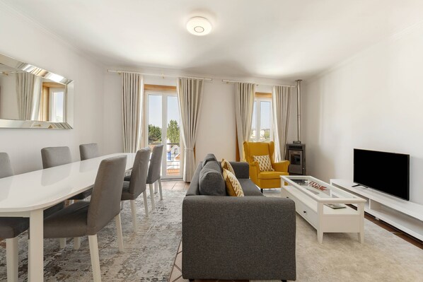 A modern living room with a dining table, sofa, TV, and a large window with curtains.