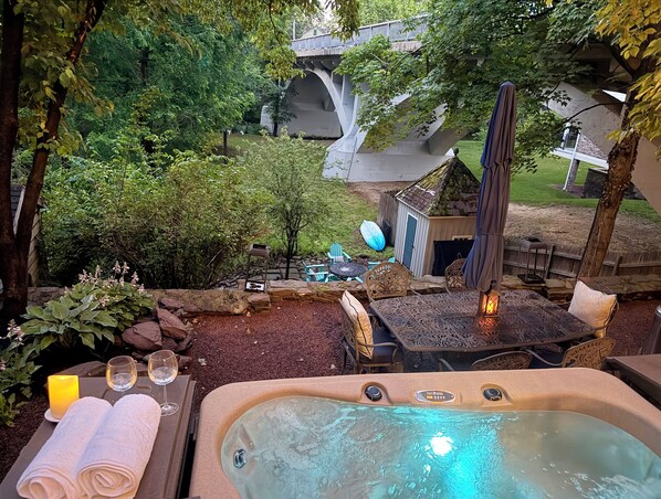 A hot tub with a control panel, a table with two glasses and towels, and a patio area with chairs and a small shed.