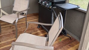 Een overdekt terras met een barbecue, twee stoelen en uitzicht op de natuur.