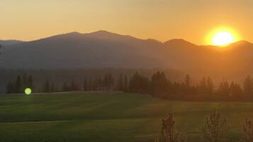 A landscape with a sun setting behind a mountain range, a field in the foreground, and trees.