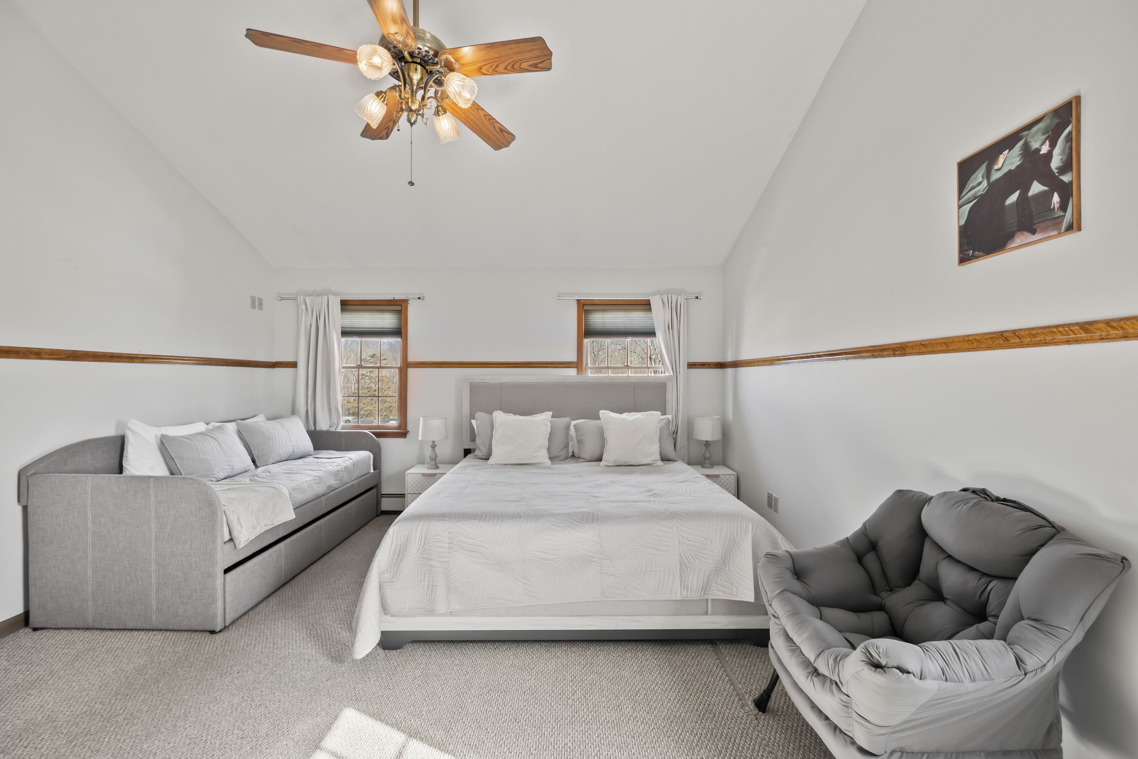 Ein Schlafzimmer mit einem Bett, einem Schlafsofa, einem Sessel, einem Deckenventilator und zwei mit Vorhängen versehenen Fenstern.