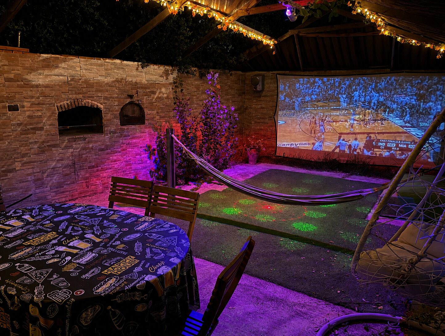 Een terras buiten, met een stenen muur, een hangmat, een tafel met een tafelkleed met patroon, en een groot scherm waarop een basketbalwedstrijd te zien is.