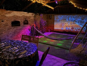An outdoor patio with a stone wall, a hammock, a table with a patterned tablecloth, and a large screen displaying a basketball game.