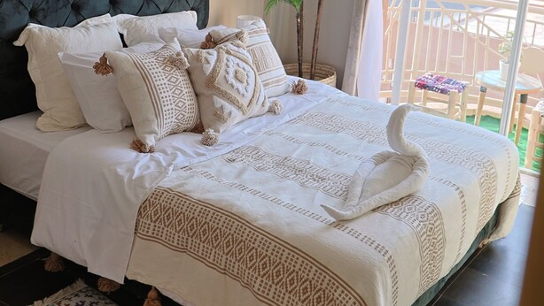 A bedroom with a tufted headboard, a bed with patterned bedding, a colorful rug, and a balcony with a view. - Appartement Moderne au Centre-Ville Guéliz (Marrakech)
