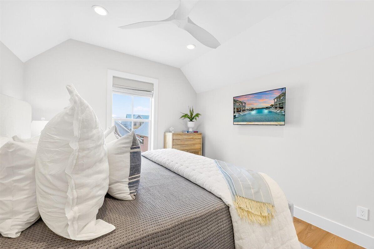 A bedroom with a bed, pillows, a plant, a painting, and a ceiling fan.