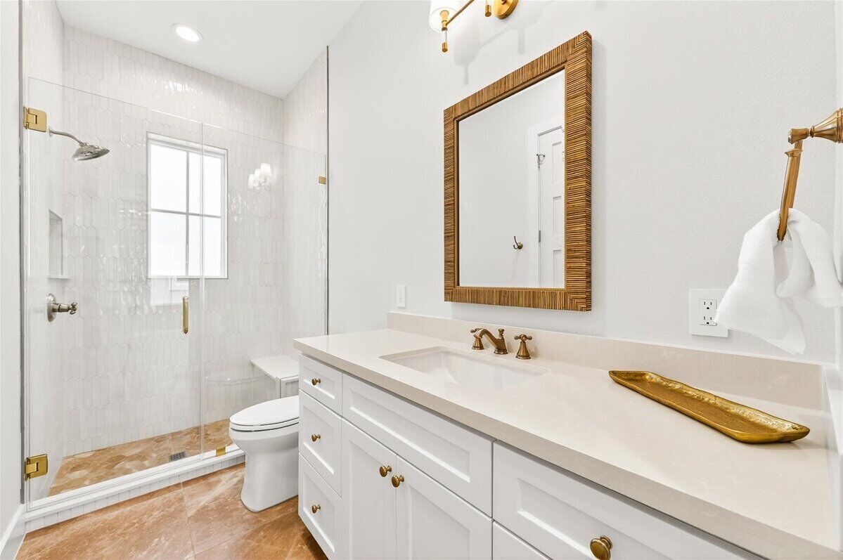 Une salle de bain moderne avec une douche à parois de verre, des toilettes blanches, un grand miroir avec un cadre doré et un lavabo blanc agrémenté de robinetterie dorée.