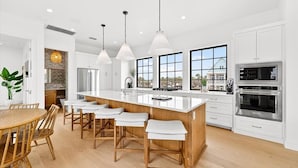 A modern kitchen with white cabinets, a large island with bar stools, stainless steel appliances, and large windows. - The Little Dipper - 100 STL (Port Aransas)