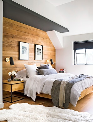 Quarto com teto inclinado, painéis de madeira, cama com várias almofadas, abajures, relógio e janela com persiana.