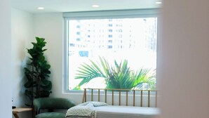 Quarto com cama, uma cadeira verde, planta em vaso e janela com vista para edifícios.