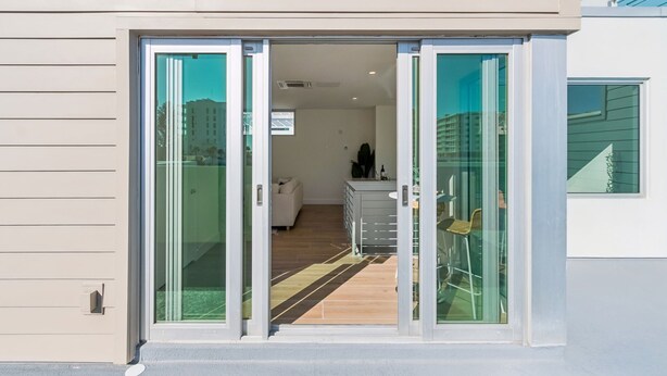 Um edifício moderno com entrada por porta de correr de vidro, interior visível com sofá e um armário, e vista desobstruída para o exterior.