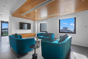 A modern living room with teal sofas, a flat-screen TV, and large windows offering a city view.