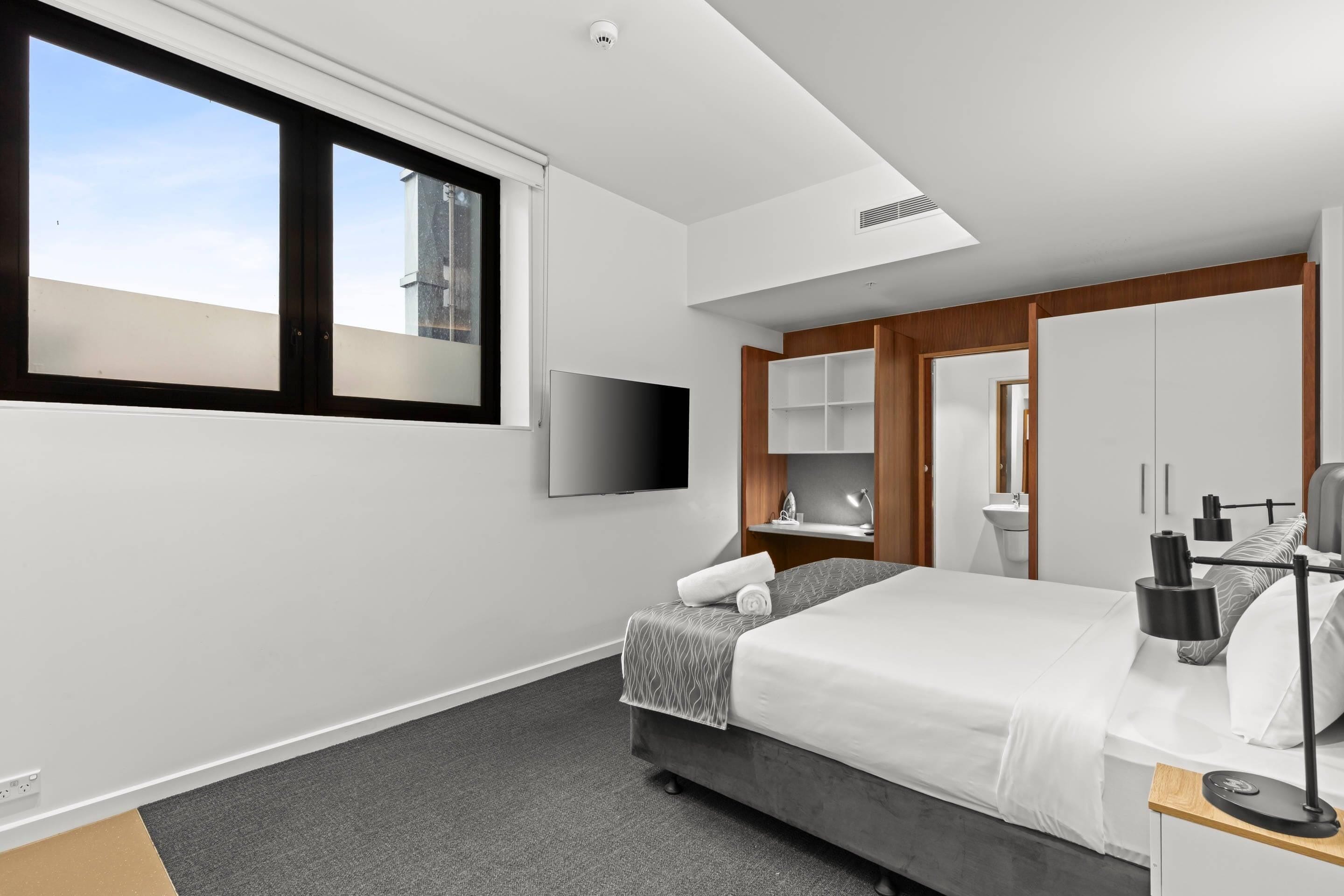 Una habitación de hotel moderna con una cama grande, un televisor de pantalla plana, un escritorio con lámpara y un baño con espejo y lavamanos.