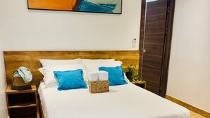 Un letto rifatto con lenzuola bianche e blu, una testiera in legno, un comodino con un cesto e una porta alla destra.