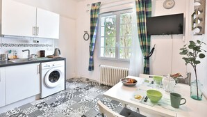 A compact kitchen with a washing machine, a dining table with bowls and mugs, a radiator, and a TV mounted on the wall. - Lavignottes - AC - Piscine & Tennis (Labouheyre)
