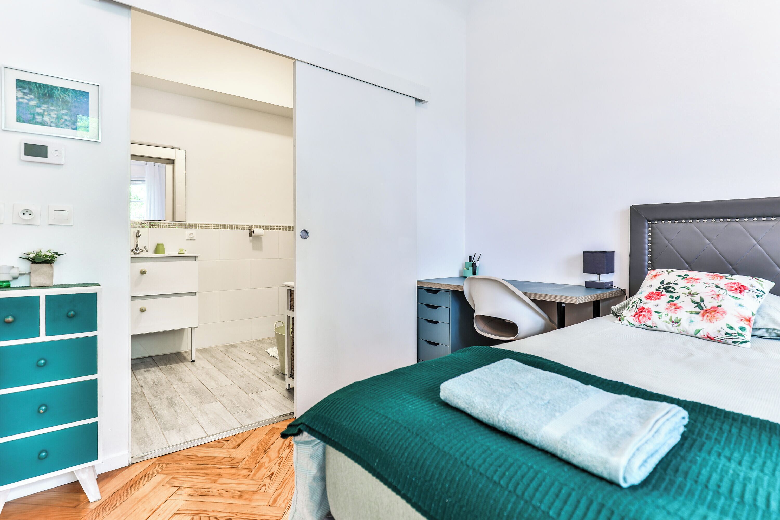 A bedroom with a bed, a desk, a chair, a wardrobe, and a bathroom visible through an open door.