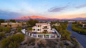 A two-story house with a pool, surrounded by desert vegetation and mountains in the background.