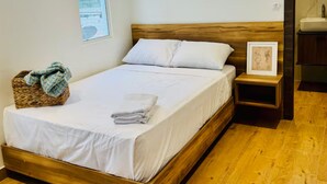 Een eenpersoonsbed met houten frame, wit beddengoed, een klein nachtkastje met een foto en een rieten mand aan de linkerkant.