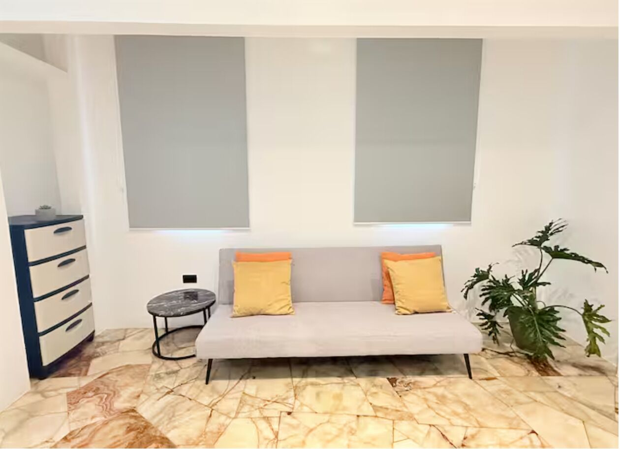 Una habitación con sofá, dos cojines, una mesita redonda, un armario y una planta en maceta.