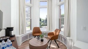 A corner room with two brown chairs, a round wooden table, and a view of the street through large windows. - The Suites | Downtown Montreal (Montreal)