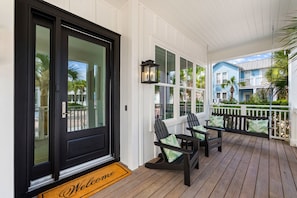 Eine überdachte Veranda mit einer schwarzen Tür, zwei Adirondack-Stühlen, einer Bank und Blick auf ein blaues Gebäude.