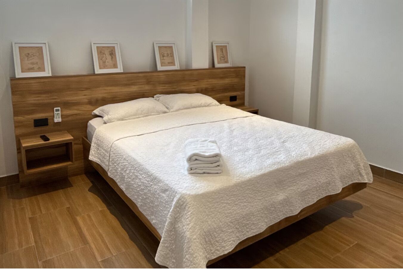 A neatly made bed with a white quilt, a wooden headboard, and a bedside table with a phone and a remote control.