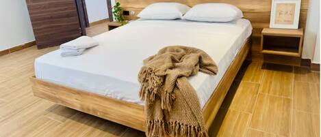 Una cama con estructura de madera, ropa de cama blanca y una manta marrón. Hay una mesita de noche con un cuadro enmarco y una planta pequeña. El piso es de madera y la puerta también.