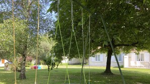 Un columpio con un asiento azul y uno amarillo, un pequeño trampolín azul, y un árbol al fondo.