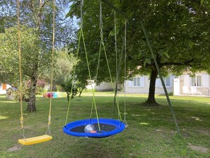 Ein Schaukelständer mit einem blauen Schaukelsitz und einem gelben Sitz, ein kleiner blauer Trampolin und ein Baum im Hintergrund.