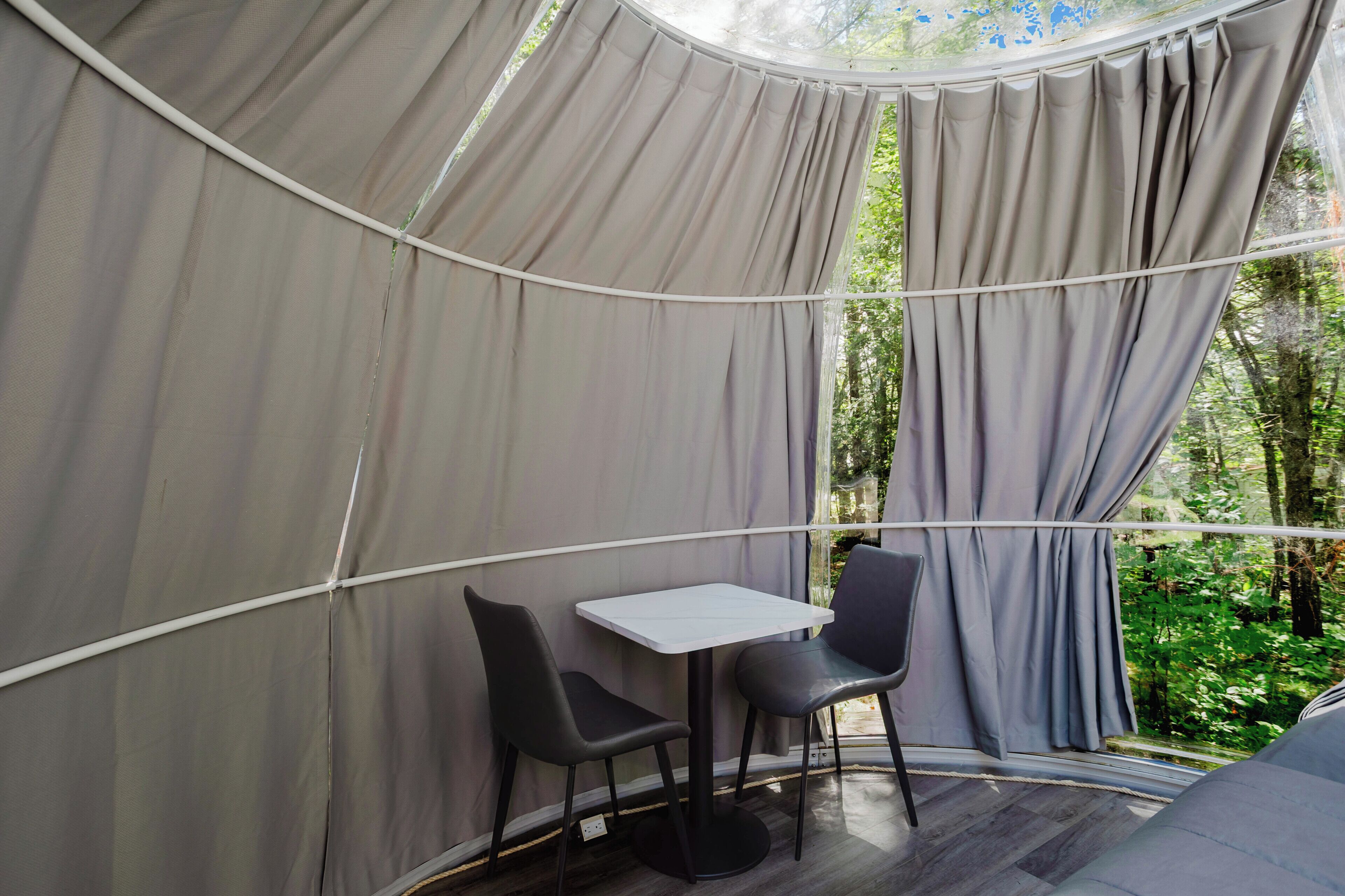 Une structure en forme de tente, avec une table et des chaises à l’intérieur, entourée de rideaux et offrant une vue sur la verdure.