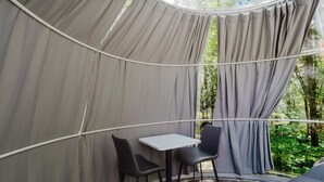 Une structure en forme de tente, avec une table et des chaises à l’intérieur, entourée de rideaux et offrant une vue sur la verdure.