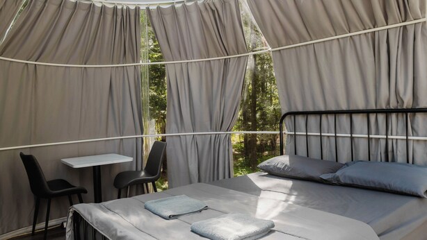 Une pièce aménagée comme une tente, avec un lit, une table et des chaises, entourée de rideaux et offrant une vue sur les arbres.