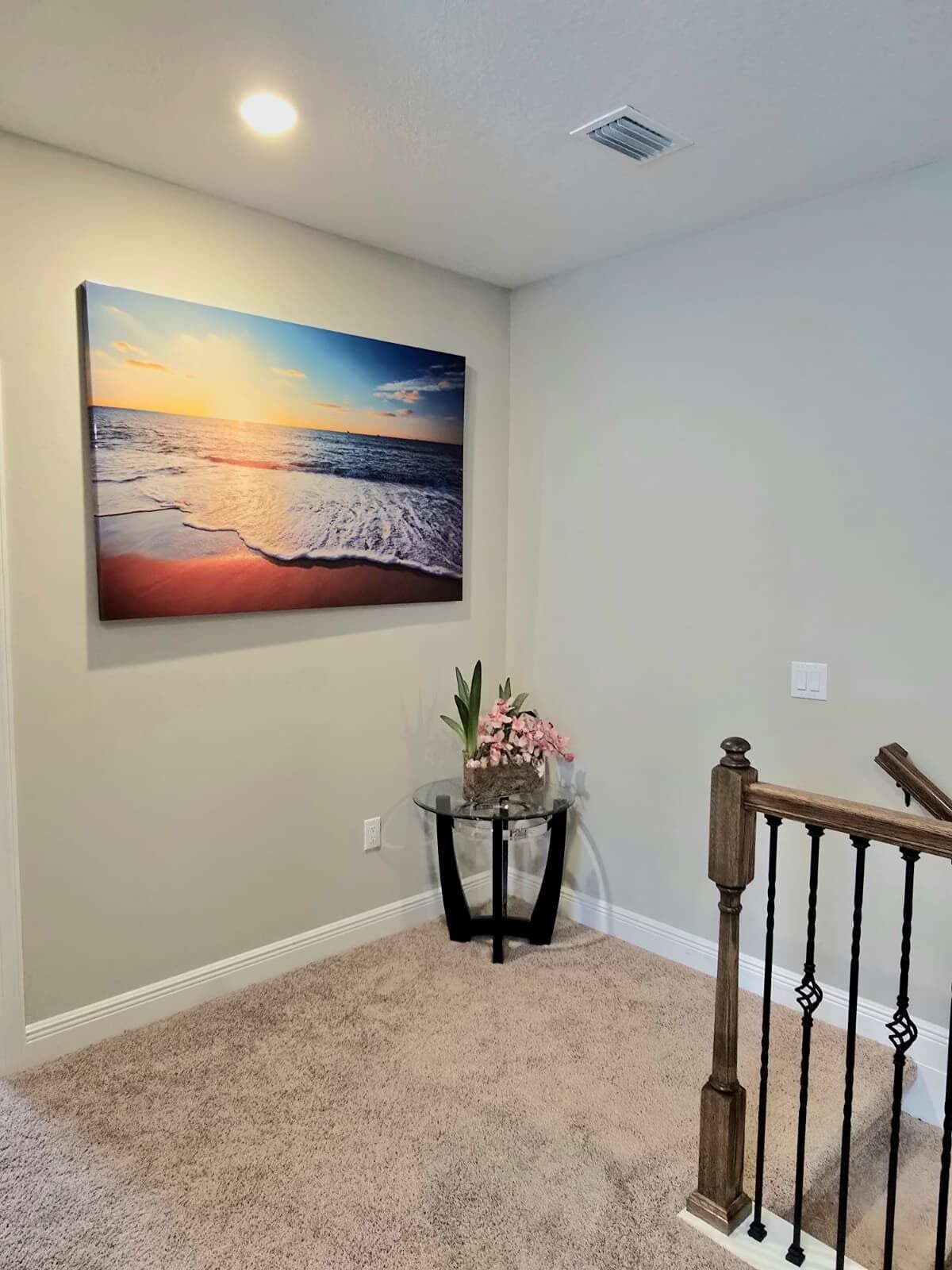 Un couloir orné d’un grand tableau représentant un coucher de soleil sur l’océan, une petite table ronde avec une plante et un escalier en bois.