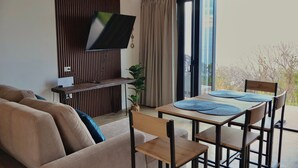 Una sala de estar con un sofá, un televisor montado en la pared, una mesa de comedor con sillas y un ventanal corredizo que da a un balcón.