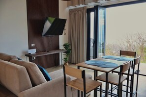 Ein Wohnzimmer mit einer Couch, einem an der Wand montierten Fernseher, einem Esstisch mit Stühlen und einer Glastür zum Balkon.