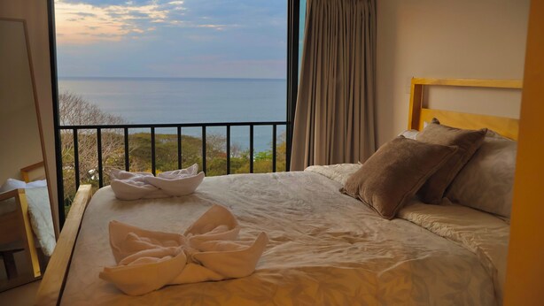 Un dormitorio con un gran ventanal, una cama con almohadas, cabecero de madera y un balcón con vistas al mar.